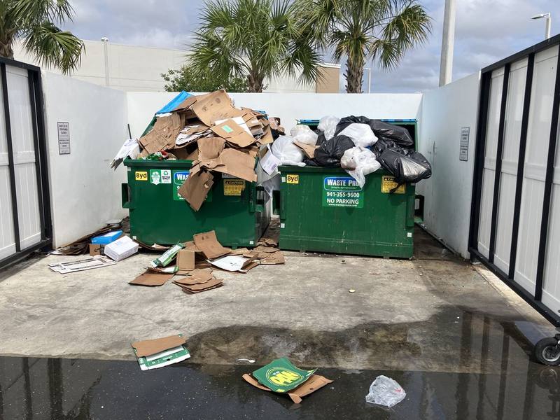 Commercial dumpster enclosure with cardboard and bags spilling onto a stained, wet concrete pad
